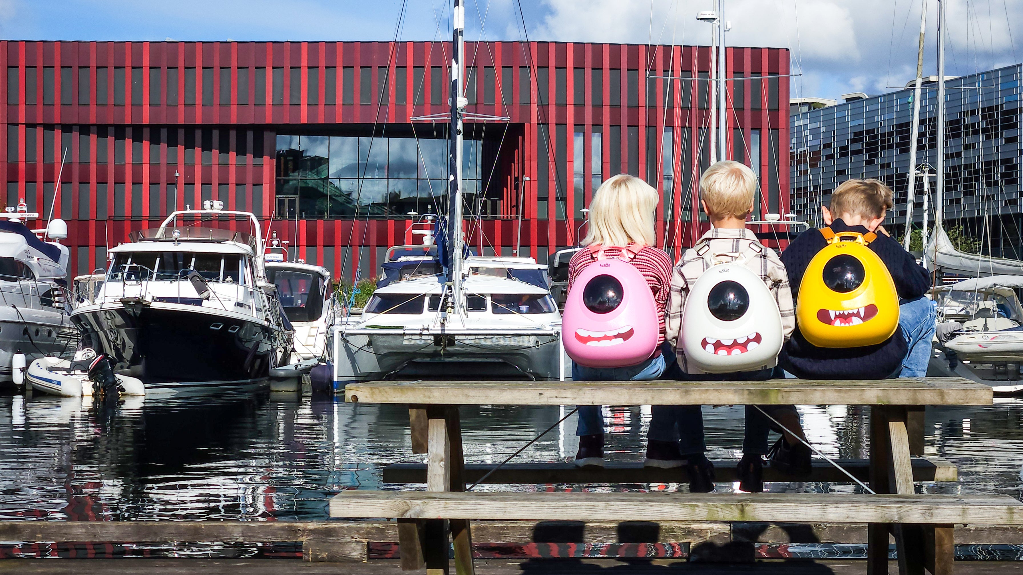 Three kids sitting on a park bench overlooking the harbor in Sandnes, wearing Ledback Buddy kids backpacks with LED screens and teeth stickers.