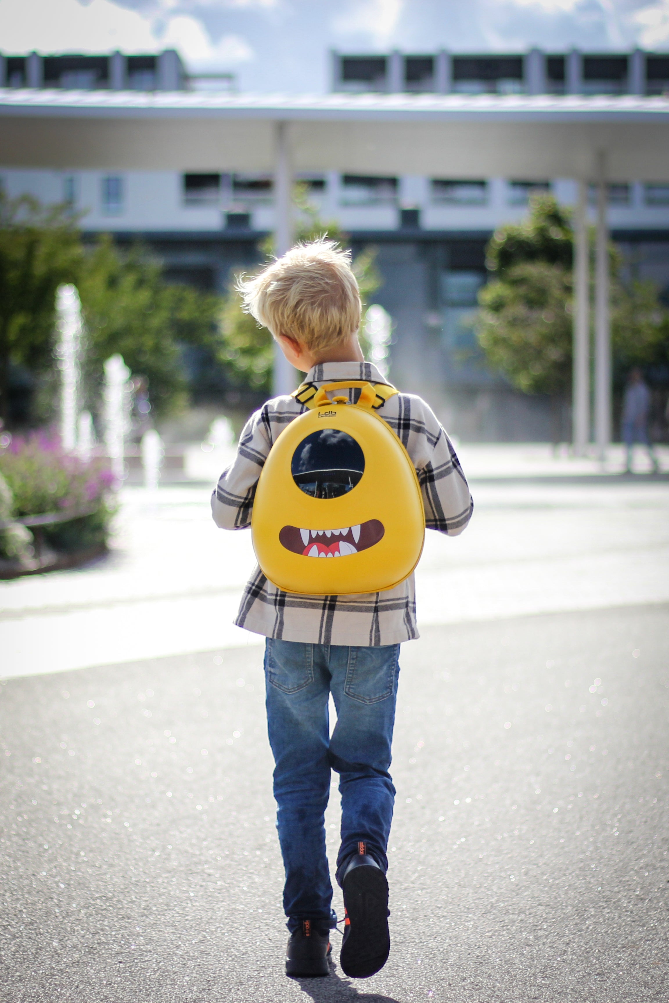 Boy walking across a sunny town square, wearing a Lemon Drop Yellow Ledback Buddy backpack with a blacked out round LED screen and a teeth sticker with "scary" pointy teeth.
