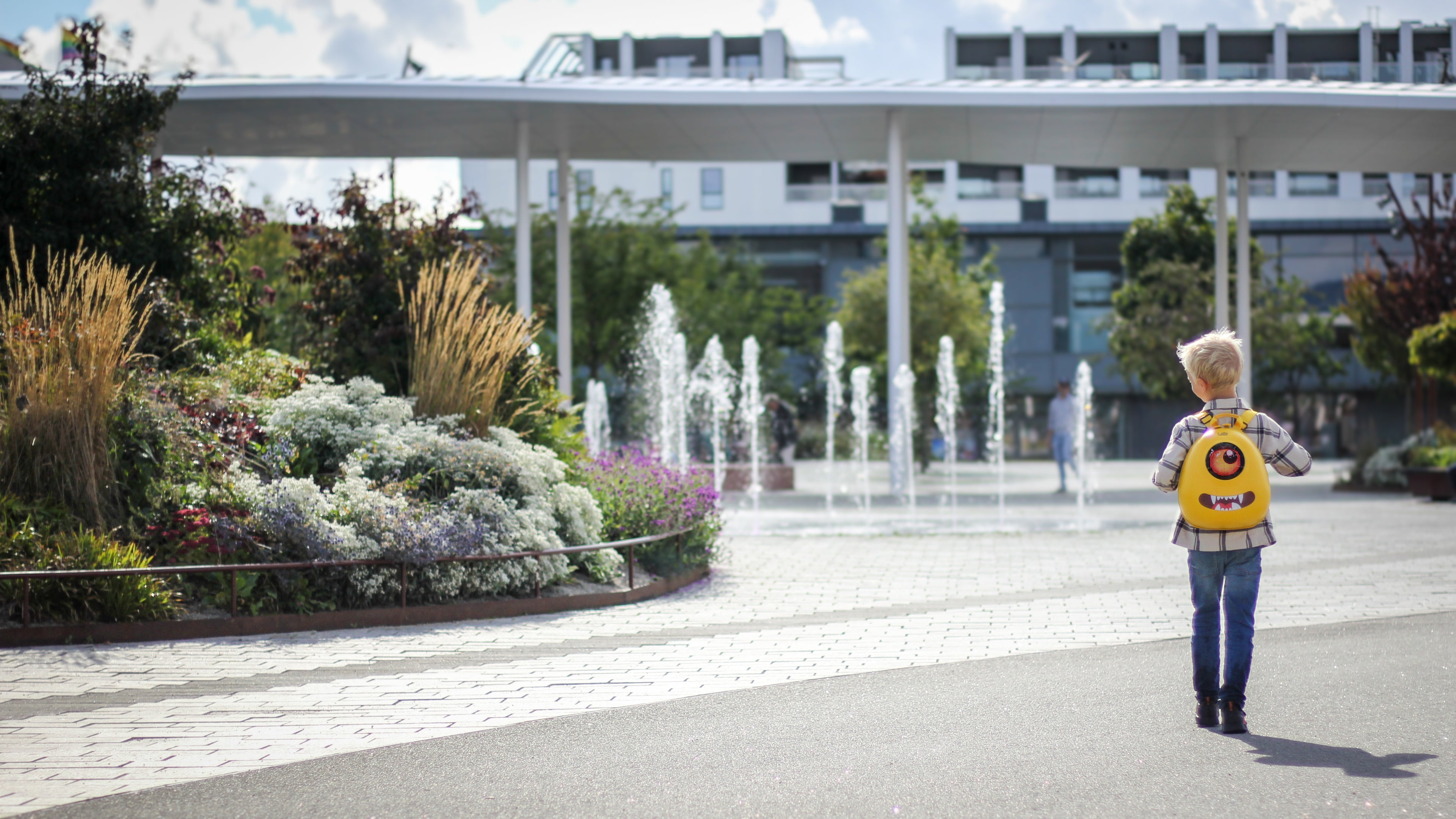 Boy walking towards a fountain in the park wearing a yellow hard shell backpack from Ledback, featuring a round LED screen displaying an orange eye and teeth sticker with pointy teeth.