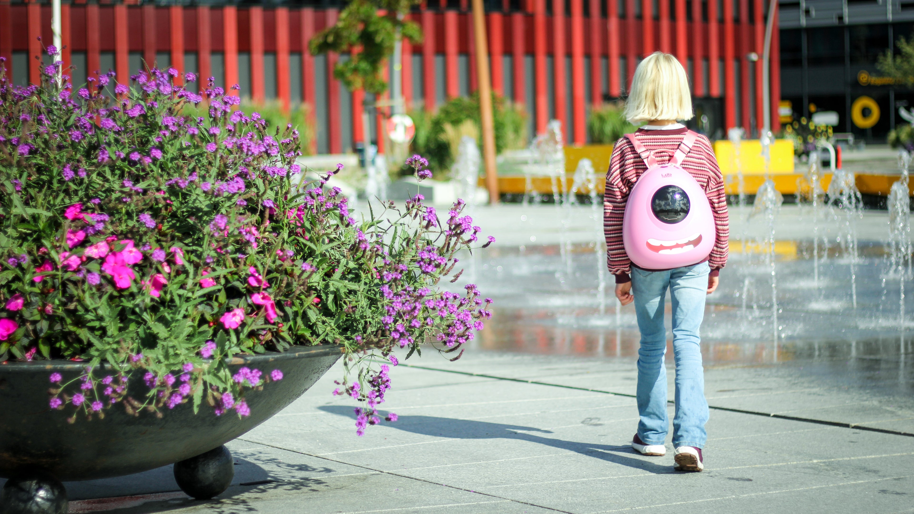 Girl in a pink sweater walking past a big pink and purple flower arrangement in a park, wearing a Raspberry Sorbet Pink Ledback Buddy backpack with an eye design LED screen and teeth sticker.