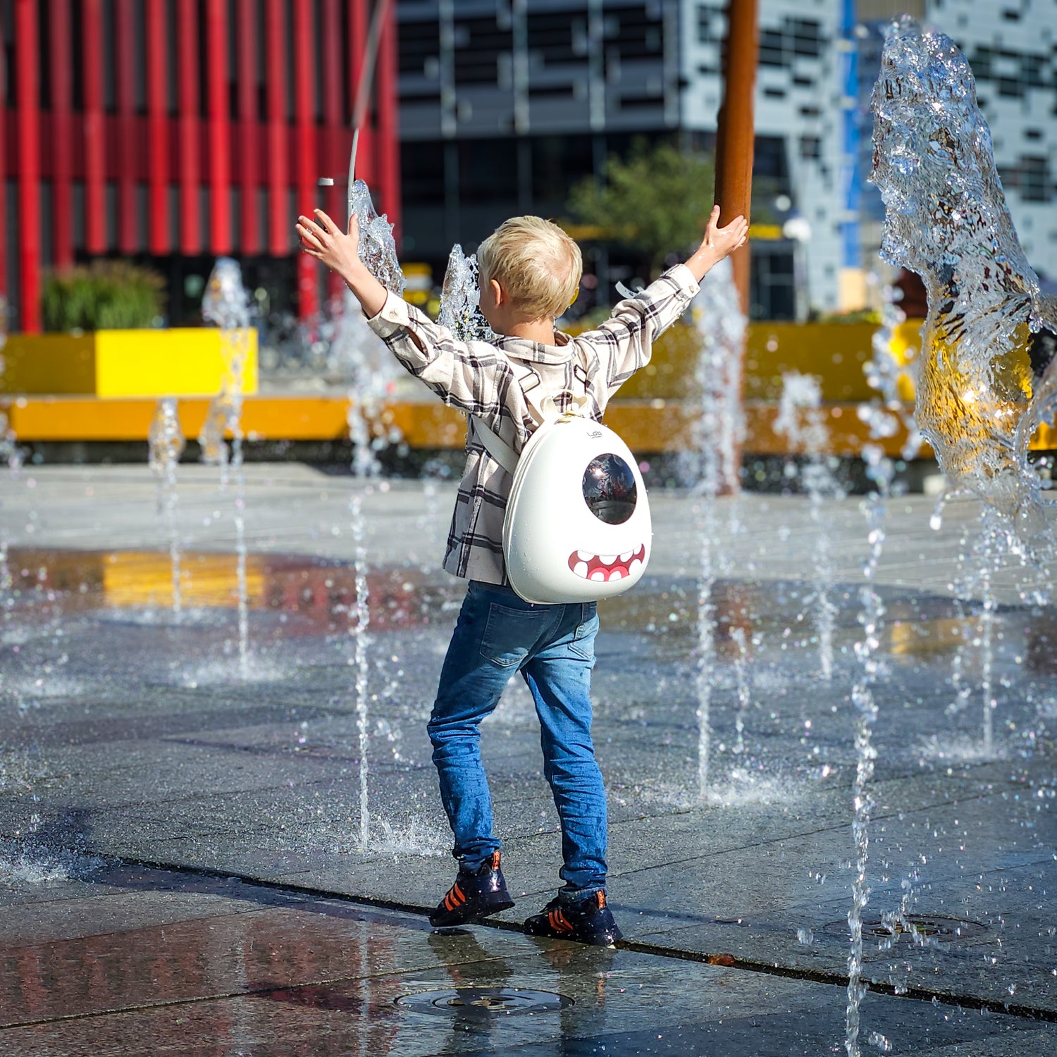 Boy wearing a Whipped Cream White hard shell Ledback Buddy backpack, featuring a round LED screen and teeth sticker, walking across a fountain with his hands stretched up in the air.