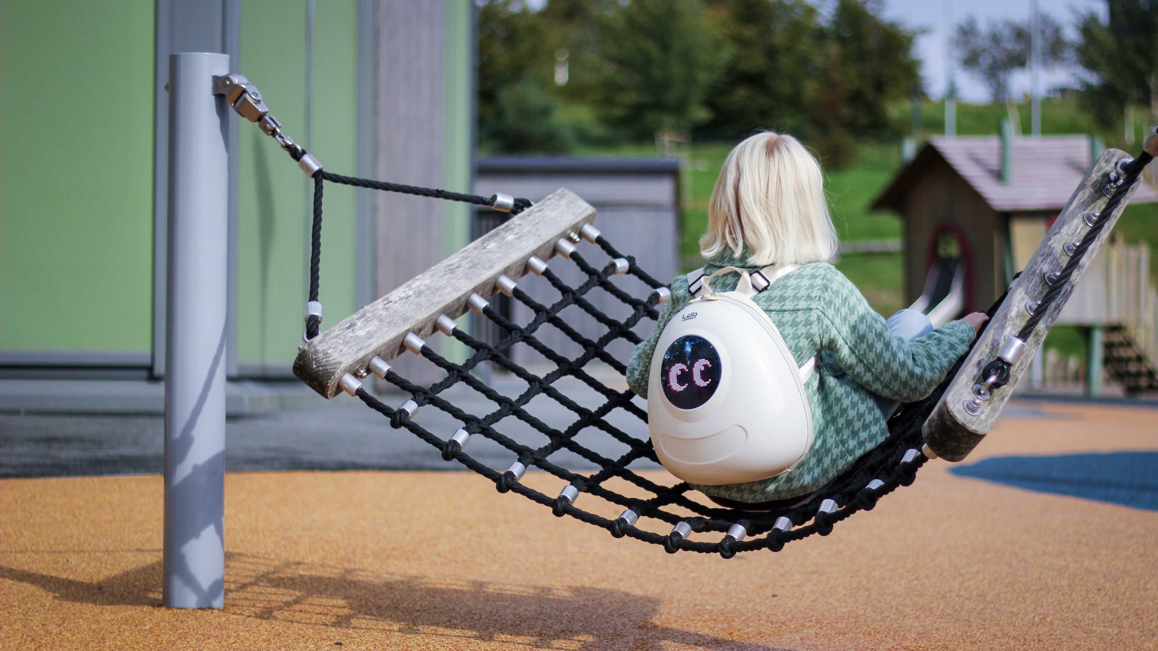 Young girl in a green jacket sitting on a hammock with a school playground in the background, wearing a Whipped Cream White Ledback Buddy backpack with an LED screen displaying two eyes looking back at you.