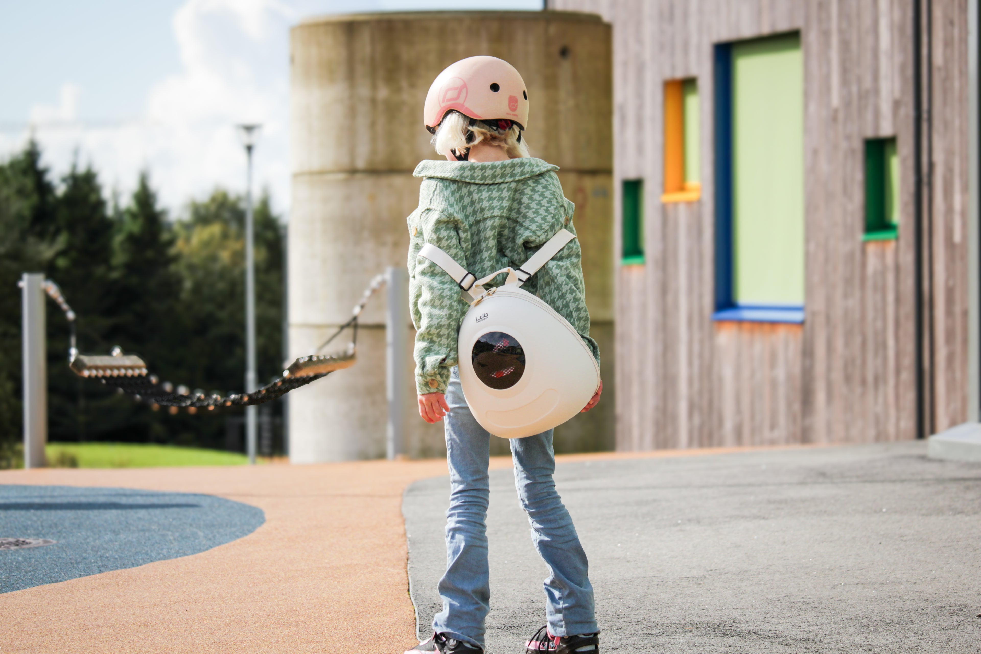 Girl on roller skates wearing a green jacket, pink helmet, taking off a Whipped Cream White kids backpack from Ledback, featuring a round LED screen with an eye design.